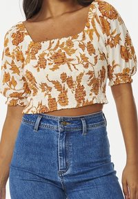 Femme portant un crop top blanc avec un imprimé floral orange et des manches bouffantes, associé à un jean en denim bleu taille haute.
