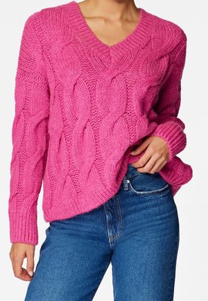 Femme portant un pull en maille épaisse rose vif à col en V avec un motif torsadé, associé à un jean bleu, main posée dans la poche.
