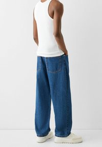 En person med vit ärmlös tröja, lösa blå jeans med bakfickor och vita sneakers, stående mot en enkel ljus bakgrund.