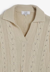 Pull en tricot beige avec un col en V et un col côtelé. Présente un motif en tricot torsadé rehaussé de petites embellissements brillants.