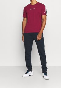 Bordeaux kortärmad t-shirt med vit "Champion"-logotyp, tillsammans med marinblå joggingbyxor med sidostripes och vita sneakers.