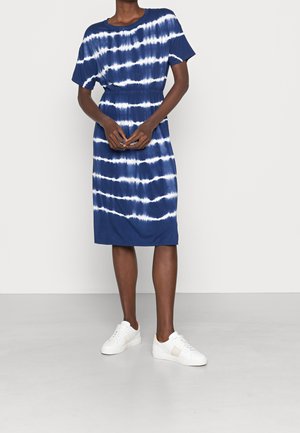 Osoba ubrana w niebiesko-białą sukienkę farbowaną na sucho, sięgającą do kolan, z krótkimi rękawami i białymi trampkami, stojąca na tle jednolitego tła.