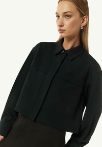 Czarna krótka koszula zapinana na guziki z dwiema dużymi kieszeniami z przodu, długimi rękawami i gładką teksturą. Posiada klasyczny kołnierzyk i dyskretny detal z logo.