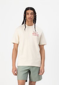 T-shirt beige a maniche corte in cotone, con testo stampato rosso. Abbinato a pantaloni corti verde chiaro, il modello si trova di fronte a uno sfondo bianco.