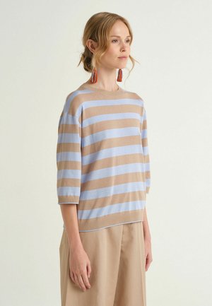 Donna con capelli raccolti che indossa un maglione a righe beige e azzurro chiaro e pantaloni beige, rivolta verso destra su sfondo semplice.