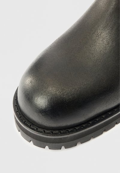 Bota de couro preta com ponta arredondada, superfície texturizada e sola de borracha robusta com padrão de aderência em zigue-zague e detalhes costurados.