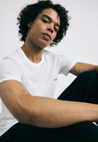T-shirt en coton blanc avec un col rond et des manches courtes, arborant un petit logo rouge sur la poitrine. Modèle assis avec les bras croisés.