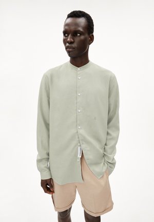 Lichtgroen overhemd met lange mouwen en een opstaande kraag, witte knopen en een geborduurd logo. In combinatie met lichtbeige korte broek.