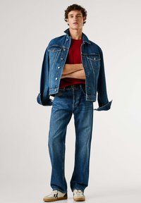 Chaqueta vaquera con dos bolsillos delanteros, de color azul, combinada con vaqueros azules y una camiseta roja. Zapatillas blancas con detalles negros.