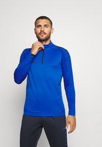 Blauw sportshirt met lange mouwen, hoge kraag, rits en textuur mouwen. Bevat zwarte accenten en logo op de borst en schouder.