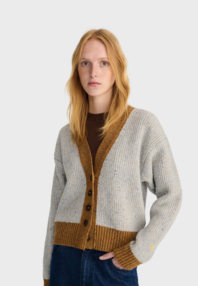 Jeune femme aux longs cheveux roux portant un cardigan gris et marron boutonné et un jean foncé, debout avec une main dans la poche sur un fond uni.