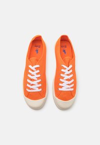 Orange canvas sneakers med vita snören, rund tå och gummisula med horisontella ribbor. Logotyp tryckt i blått på innersulan.