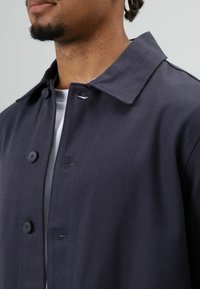 Donkerblauwe jas met knopen, met een spreidkraag, gestructureerde stof en zichtbare stikseldetails langs de voorkant.