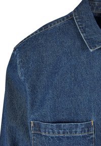 Denim krekls ar apkakli, kuram ir krūšu kabata. Spēcīga zila krāsa, teksturēts audums un redzamas šuves akcenti gar malu.