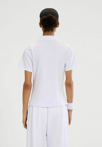 Persona con cabello rizado oscuro recogido en un moño, vistiendo una camiseta polo blanca de manga corta, pulsera blanca y pantalones blancos, de espaldas a la cámara.