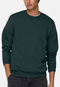 Sweatshirt vert foncé, col rond, poignets et ourlet côtelés, texture douce, coupe décontractée. Main dans la poche, porté avec un pantalon gris foncé.
