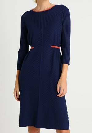 Femme portant une robe en maille côtelée bleu marine avec des manches trois-quarts et une bordure orange au niveau de l'encolure et de la taille.