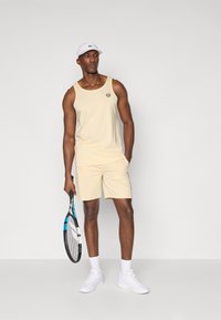 Beige tank top, kerek logóval, hozzá illő bézs sorttal és fehér tornacipővel. Kék díszítésű teniszütőt tart a kezében. Egyszerű háttér.