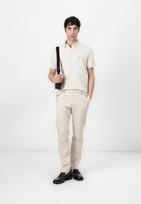 Lichtbeige poloshirt met een logo, gecombineerd met beige broek en zwarte leren schoenen. Het model houdt een zwarte schoudertas vast.