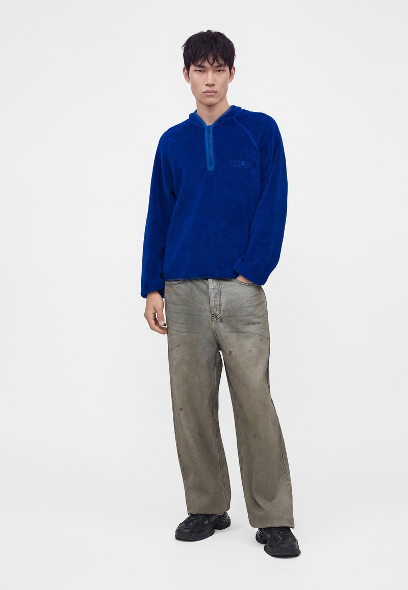 Young man standing wearing a blue fleece pullover with a front zipper and loose gray pants with black shoes against a white background.