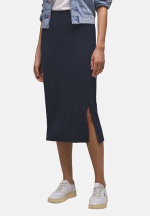 Femme portant une jupe midi bleu marine avec une fente latérale, des baskets blanches, un haut blanc et une veste en jean courte, debout sur un fond uni.