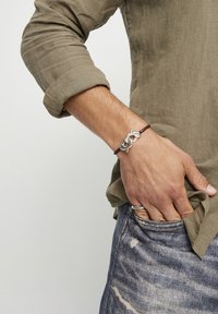 Bracelet en argent avec un design entrelac�é, attaché à un cordon marron, porté à la main. La chemise est de couleur vert olive, avec les manches retroussées.