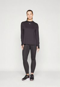 Zwart sportief langwerpig shirt met ronde hals en Puma-logo, gecombineerd met zwarte leggings en zwarte sportschoenen met roze accenten.
