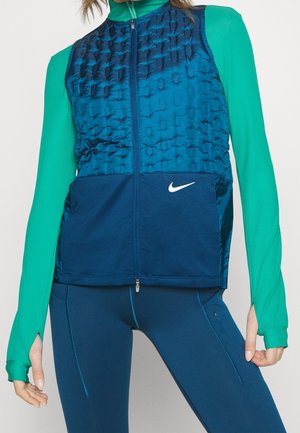 Person iført blå quiltet Nike vest over en grønblå langærmet top med tommelfingerhul og matchende blå leggings.