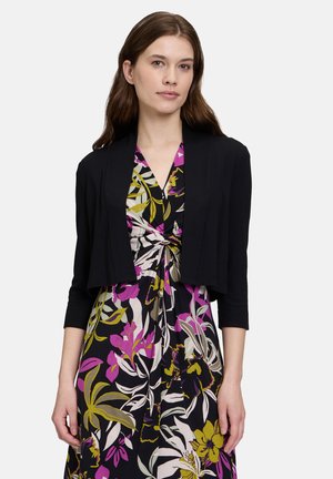 Une femme aux longs cheveux bruns porte un cardigan noir court par-dessus une robe à motifs floraux comportant des nuances de violet, jaune et vert.