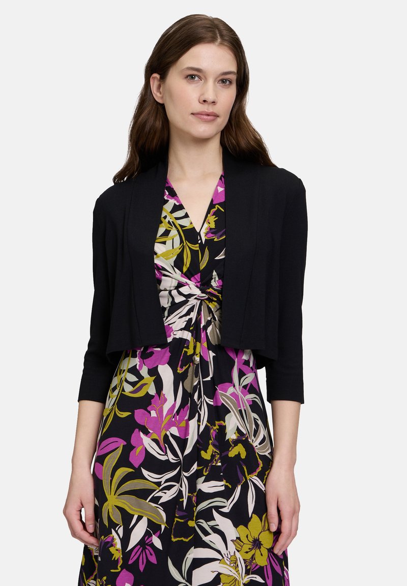 Een vrouw met lang bruin haar draagt een zwarte cropped cardigan over een bloemenjurk met paarse, gele en groene patronen.