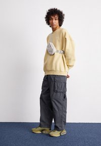 Ljusgul sweatshirt, grå cargobyxor, olivgröna sneakers med bubbel-liknande detaljer och en grå axelväska. Avslappnad och funktionell klädsel.