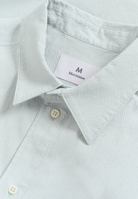 Hellgraues Hemd mit strukturiertem Stoff, spitzem Kragen und zwei Knöpfen. Das Etikett mit der Aufschrift "Matinique" befindet sich am inneren Kragen.