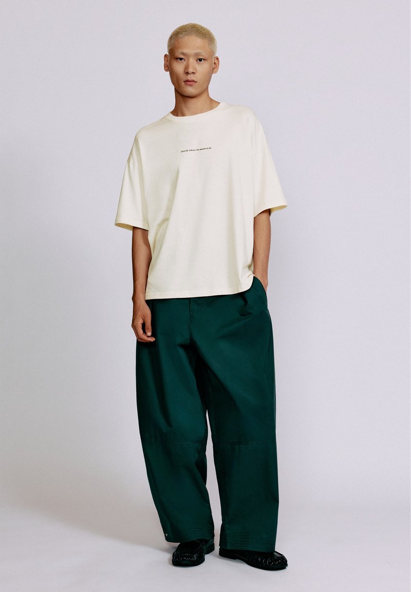 Jeune personne aux cheveux blonds courts portant un t-shirt oversize blanc cassé et un pantalon large vert foncé, debout devant un fond uni.