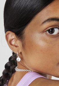 Pendientes de aro de plata con colgantes circulares texturizados, llevados con un collar metálico, sobre un fondo de piel suave y ligeramente bronceada.