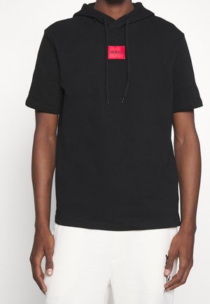 Mężczyzna w czarnej krótkiej bluzie z kapturem z czerwonym naszywką z logo HUGO, w białych spodniach, stoi na jednolitym tle.