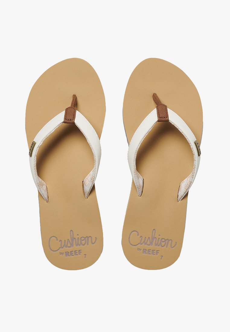 Hvite flip-flops med en brun accentstropp, med en teksturert tan såle. Præget "Cushion by REEF" tekst i lys lilla.