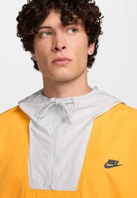 Cortavientos con capucha amarillo y gris, con media cremallera en el frente, capucha con cordón elástico y logo azul de Nike en el pecho izquierdo. Textura suave.