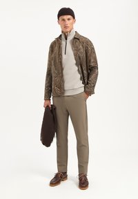 Bronzen jas met paisley-patroon over een lichtgrijze zip-up, gecombineerd met beige broek en bruine schoenen, met een bruine sjaal in de hand.