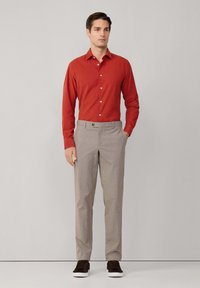 Camisa roja de manga larga con botones, pantalones grises con estampado, zapatillas oscuras, postura de pie, fondo liso.
