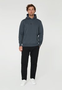 Mörkgrå hoodie med ficka fram och dragsko på huvan, prydd med en logotyp på bröstet, tillsammans med svarta byxor och vita sneakers.