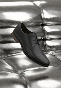Zapato de vestir de cuero negro pulido con cordones, reposando sobre una superficie acolchada brillante de color plateado metálico.