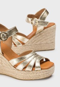 Sandales à talon en métal doré avec larges brides et fermeture à la cheville. Semelle en jute tressé avec finition texturée, design à bout ouvert.