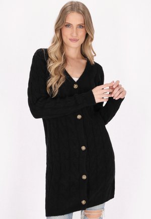 Schwarze Strickjacke mit V-Ausschnitt, langen Ärmeln, fünf braunen Knöpfen auf der Vorderseite und lockerer Passform, kombiniert mit zerrissenen Jeans.