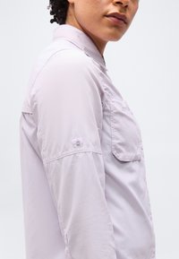 Κοντινό πλάνο ενός ατόμου που φοράει ανοιχτό μωβ πουκάμισο με μακριά μανίκια, που έχει τσέπη στο στήθος και τα μανίκια του είναι γυρισμένα και στερεωμένα με κουμπί.