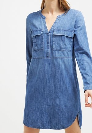 Mujer con vestido largo de mezclilla azul de manga larga con bolsillos en el pecho y una abertura frontal en el dobladillo, de pie sobre un fondo blanco liso.