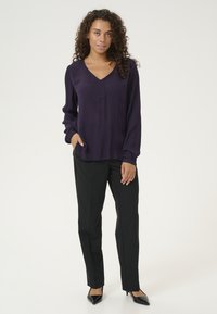 Blusa lunga viola con scollo a V e una sola tasca, abbinata a pantaloni neri sartoriali e décolleté neri a punta.