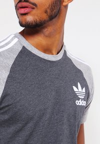 Joven con barba corta que lleva una camiseta gris de Adidas con logo y rayas blancas, mirando hacia un lado.