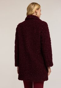 Burgundská umělá kožešina s texturovaným, plyšovým povrchem. Má široký límec a volný střih, sahající do poloviny stehna. Měkký a teplý vzhled.