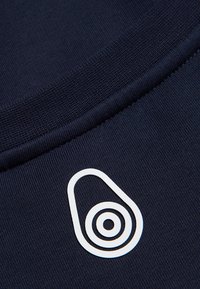 Tela azul marino con una textura suave, que presenta un logotipo gráfico blanco con forma de aguacate y círculos concéntricos.