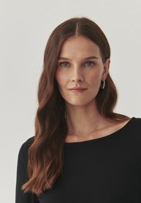 Zwarte langarmtop met een brede halslijn, gedragen door een persoon met golvend bruin haar. Bevat zilveren oorbellen in de vorm van een ring en heeft een gladde huidtextuur.
